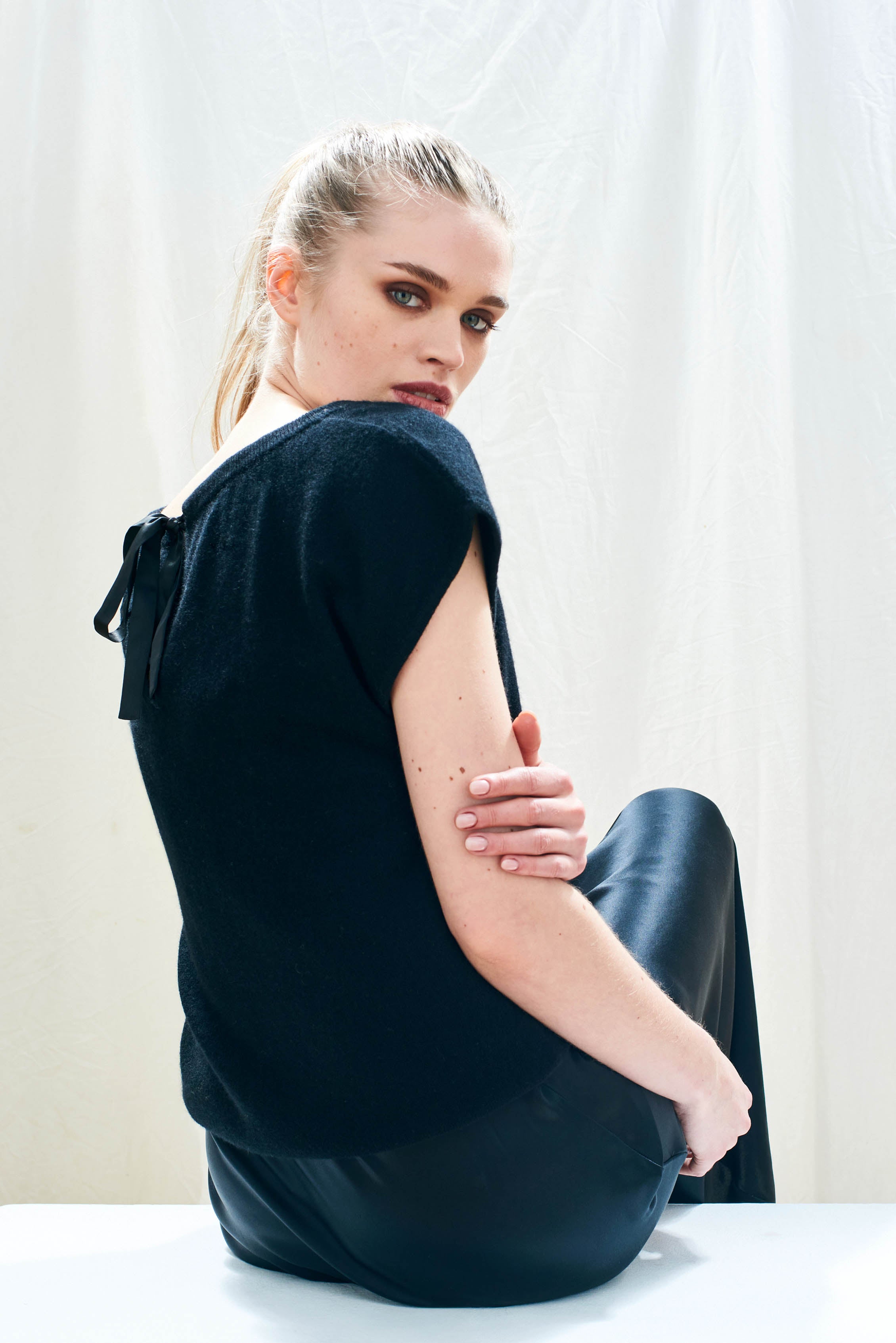 Female model wearing Jumper 1234 cap sleeve black cashmere crew neck jumper with ribbon tie detail at the back facing away from the camera