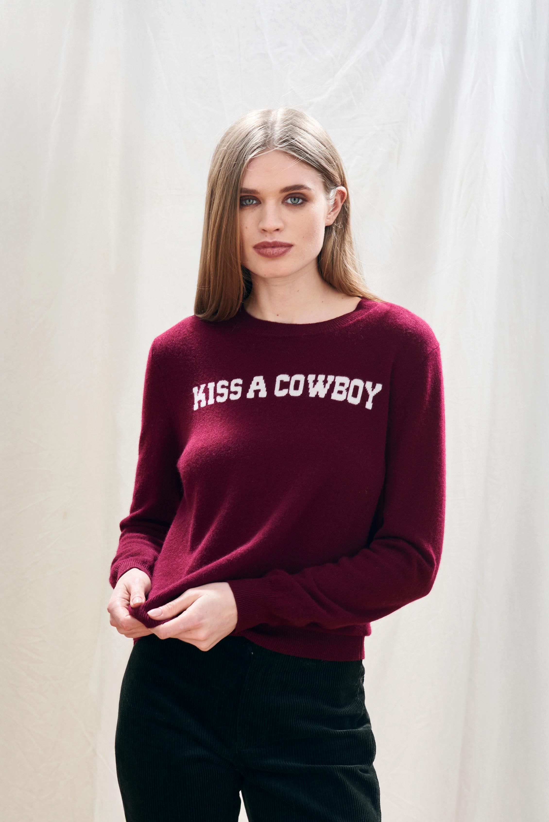 Blonde female model wearing burgundy cashmere/wool jumper with 'KISS A COWBOY' cream text intarsia