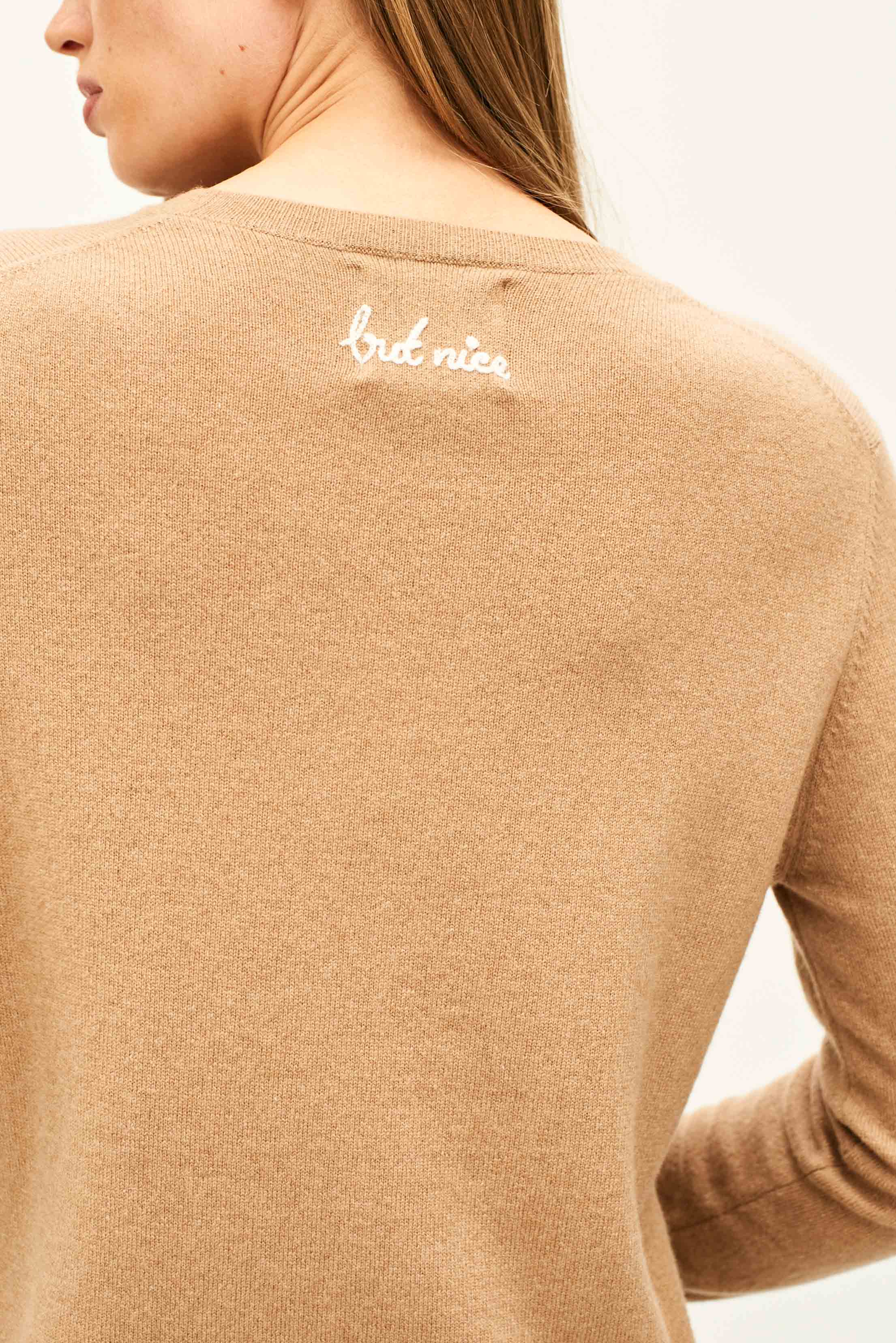 Female model wearing Jumper 1234 Camel cashmere crew neck jumper with cream 'naughty' chain stitch embroidery on the chest and 'but nice' on the back. Back shot