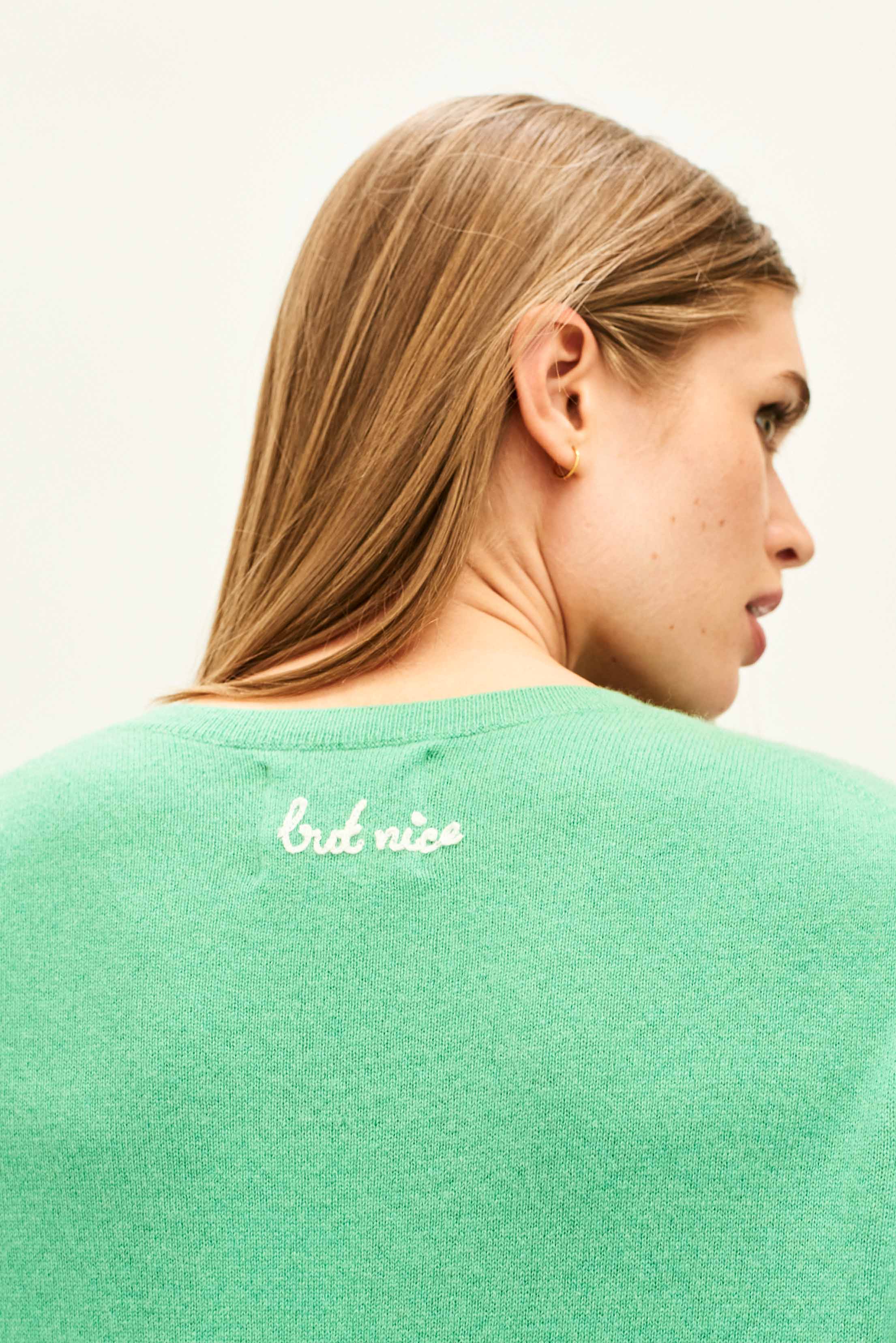 Female model wearing Jumper 1234 Green cashmere crew neck jumper with cream 'naughty' chain stitch embroidery on the chest and 'but nice' on the back. Back shot