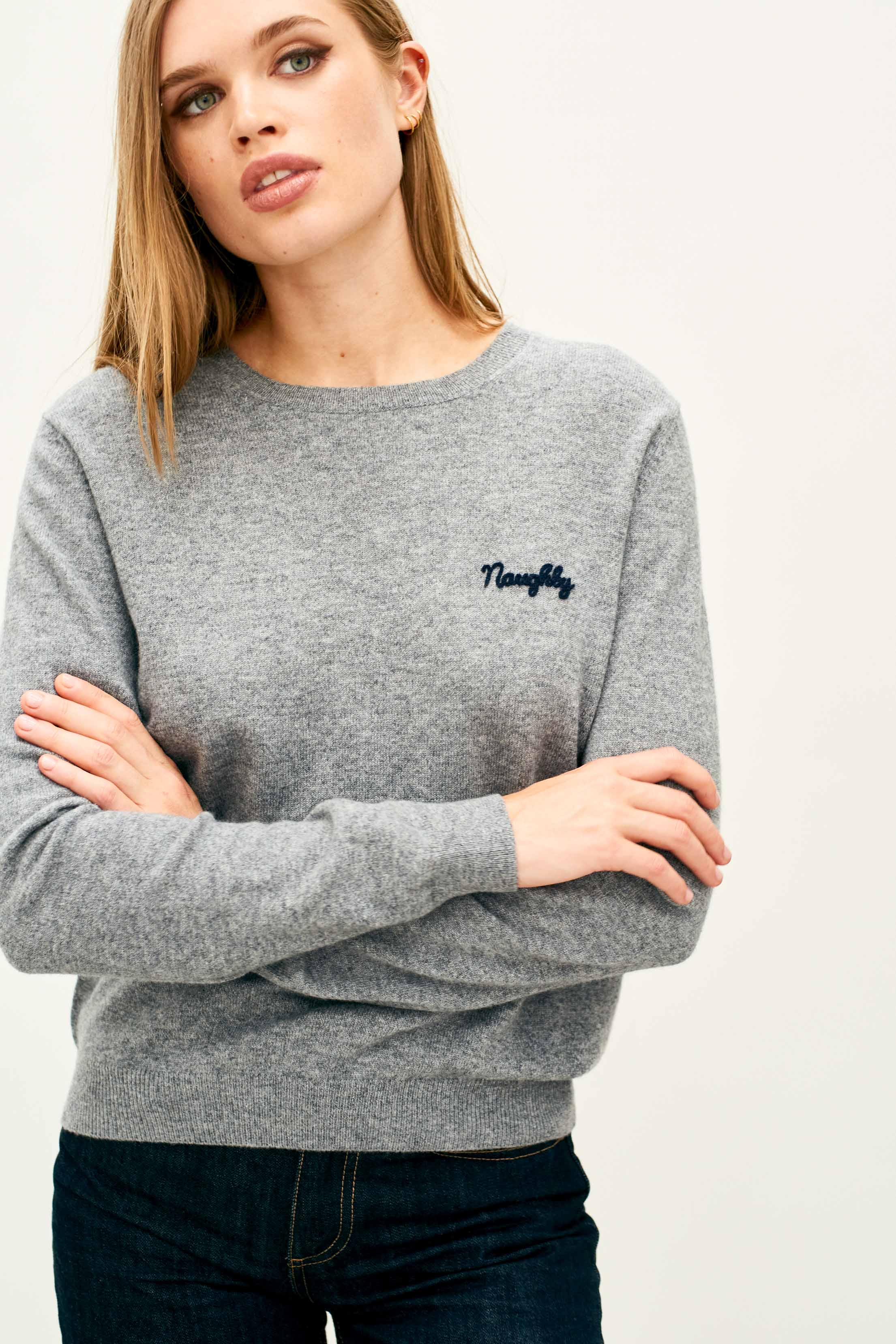 Female model wearing Jumper 1234 Mid grey cashmere crew neck jumper with navy 'naughty' chain stitch embroidery on the chest and 'but nice' on the back.