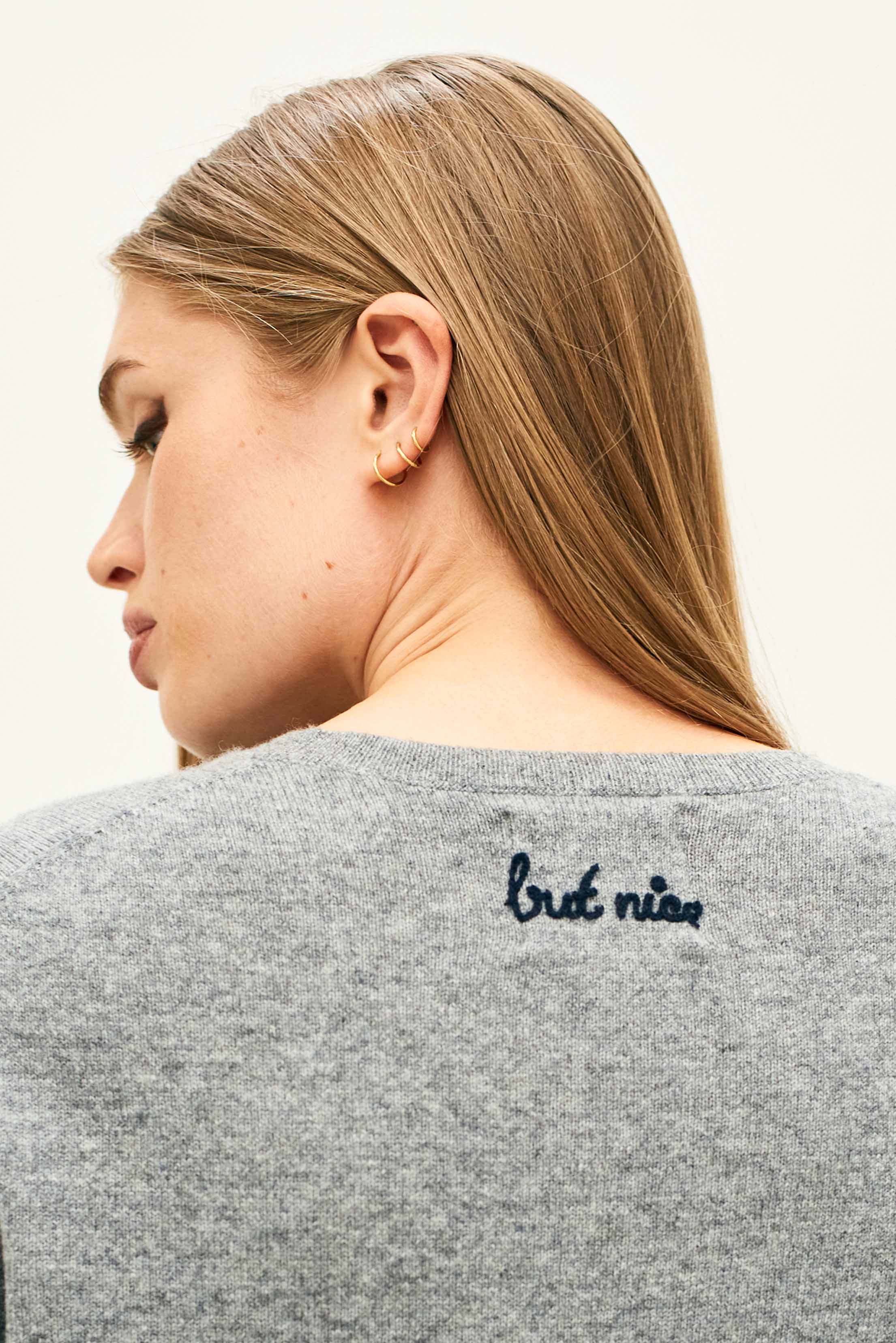 Female model wearing Jumper 1234 Mid grey cashmere crew neck jumper with navy 'naughty' chain stitch embroidery on the chest and 'but nice' on the back facing away from the camera