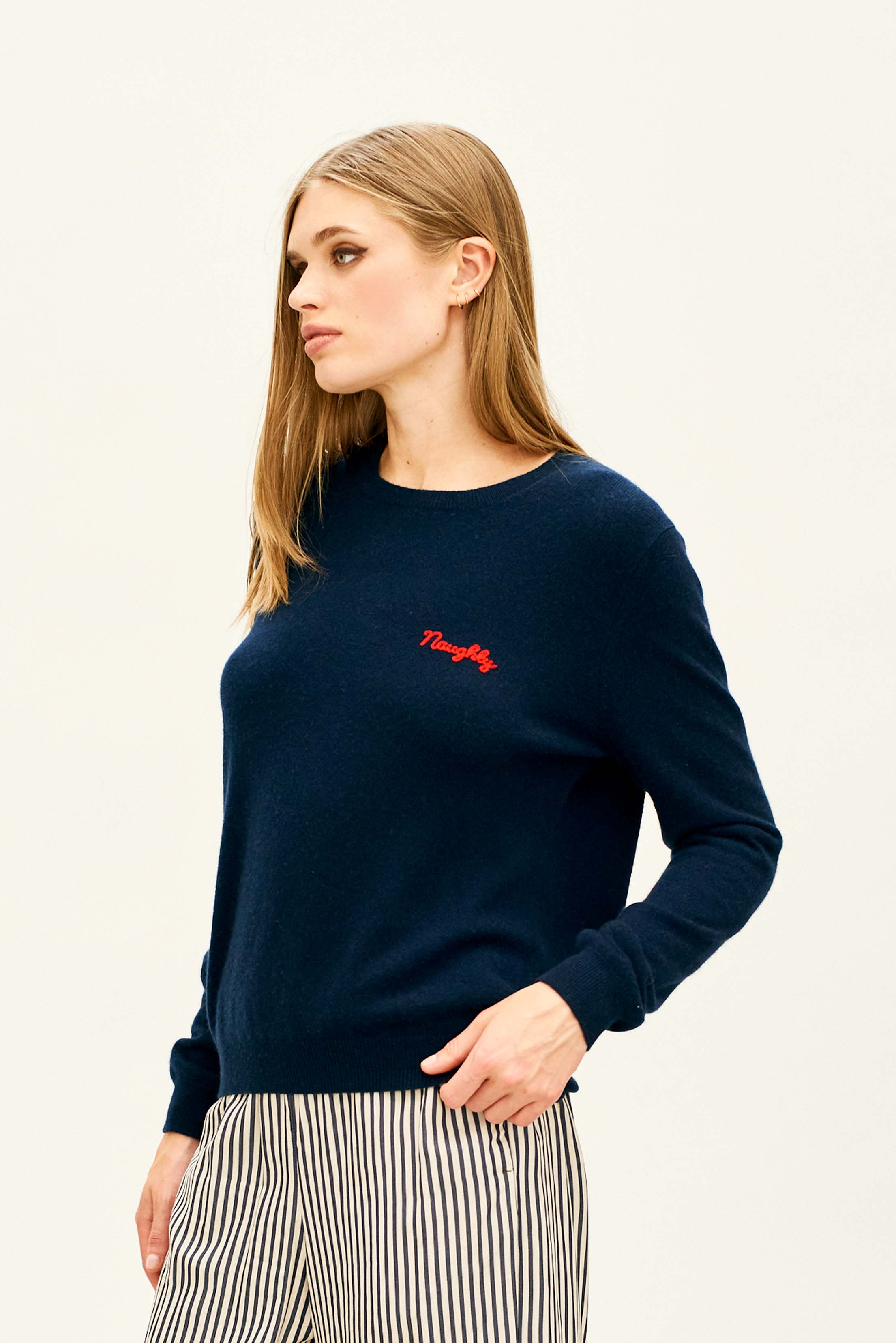 Female model wearing Jumper 1234 Navy cashmere crew neck jumper with red 'naughty' chain stitch embroidery on the chest and 'but nice' on the back.