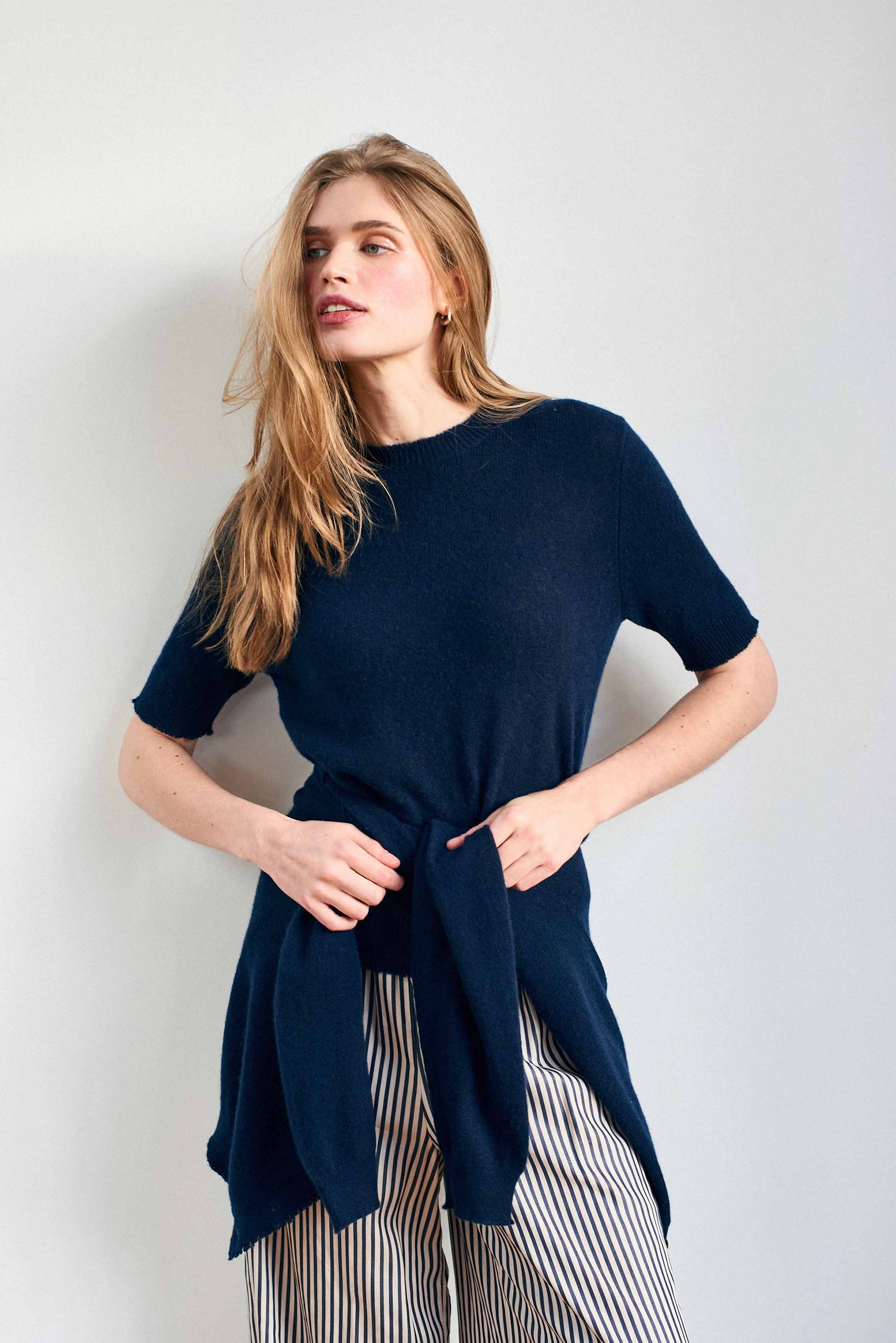 Female model wearing Jumper 1234 Navy short sleeve cashmere jumper with 'distressed' ribs. With matching cardigan toed around her waist