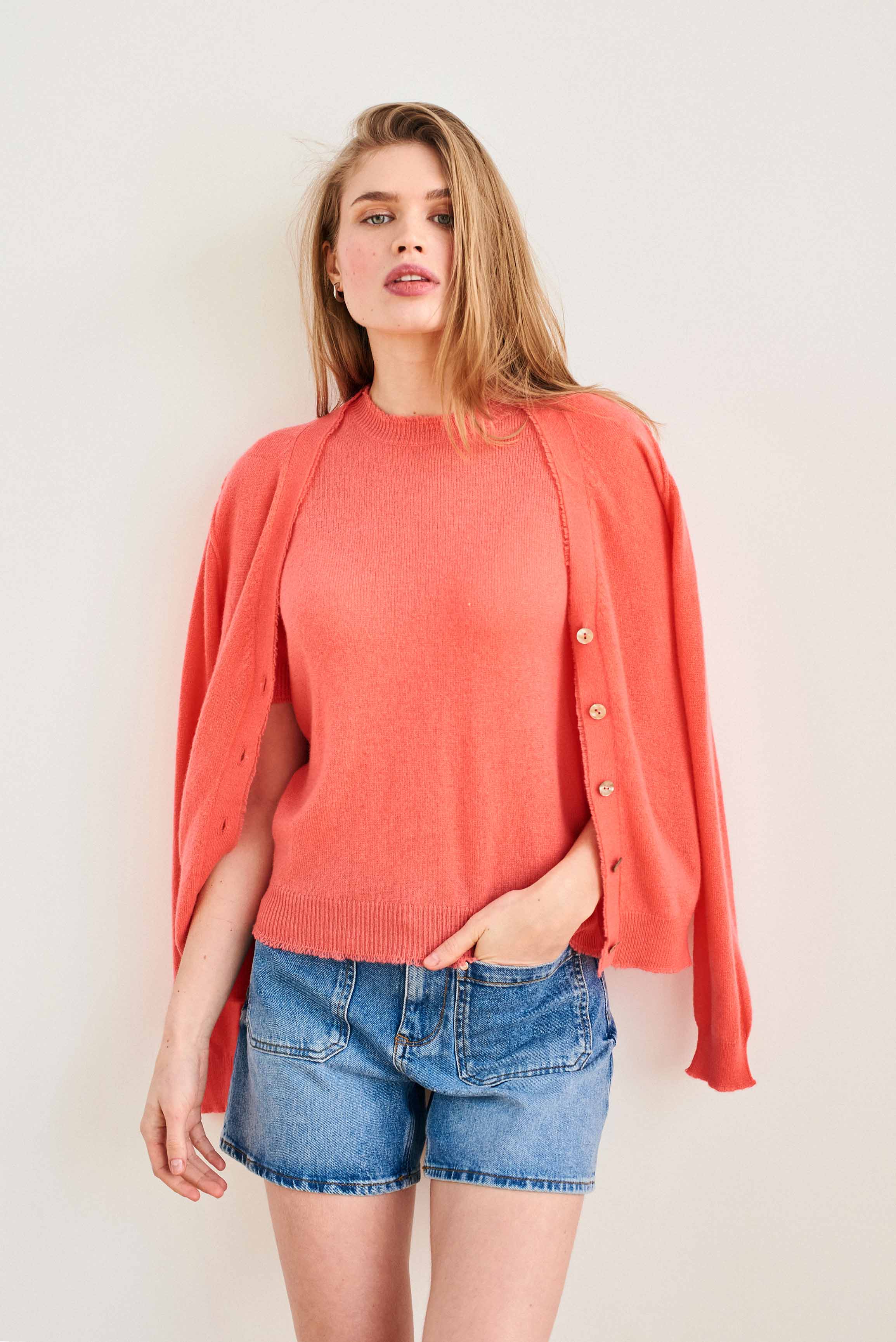 Female model wearing Jumper 1234 Coral short sleeve cashmere jumper with 'distressed' ribs. With matching cardigan draped over her shoulders