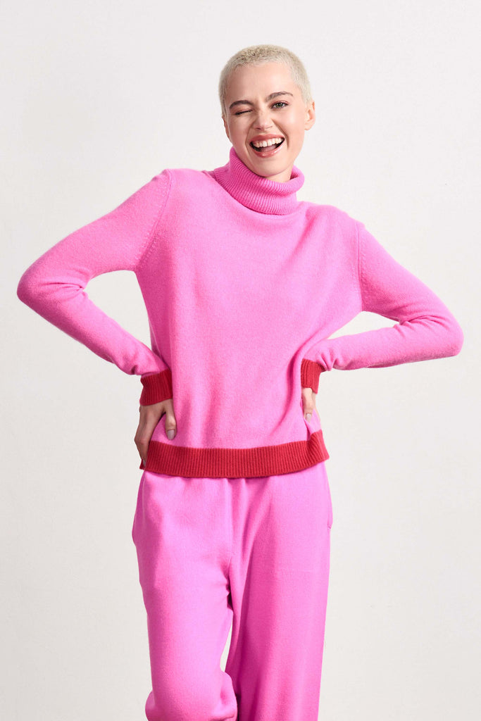 Blonde female model wearing Jumper 1234 peony pink cashmere roll neck with contrast cherry ribs.