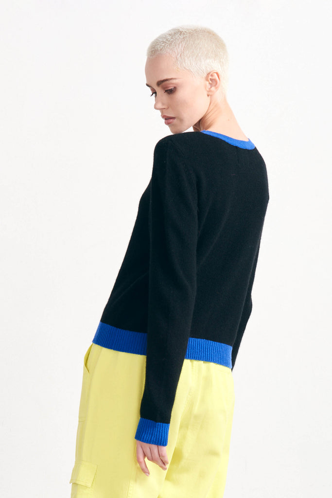 Blonde female model wearing Jumper 1234 black cashmere crew neck with contrast bright blue ribs facing away from the camera