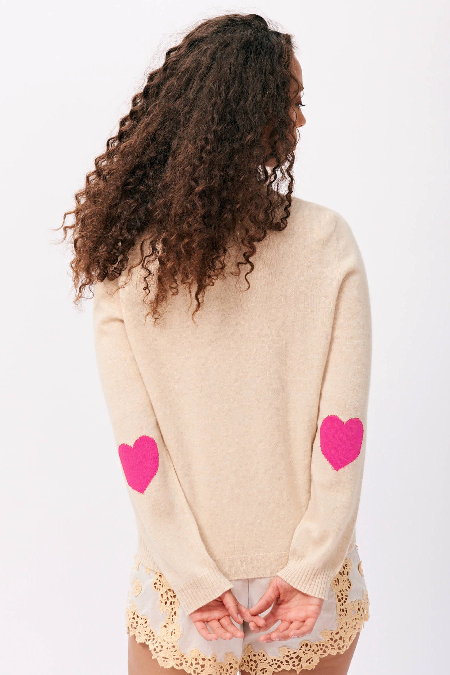 Brown haired female model wearing Jumper 1234 cashmere heart patch cardigan in oatmeal with hot pink heart elbow patches facing away from the camera