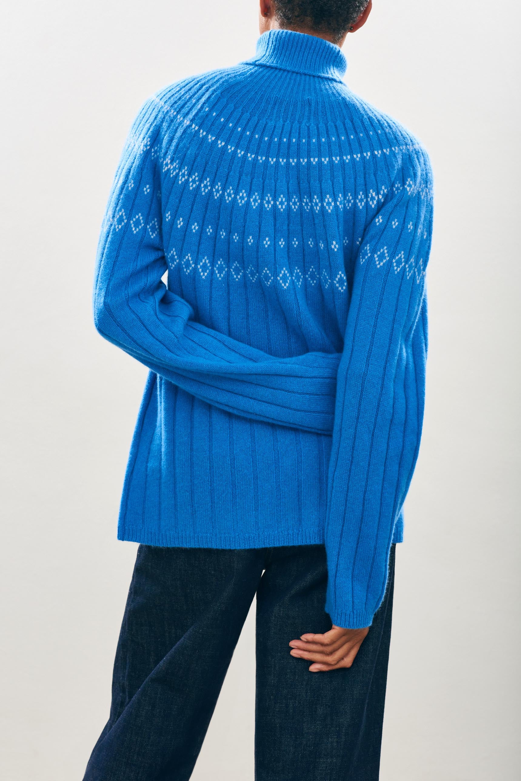 Female model wearing Jumper1234 Cashmere Rib Ski Roll Collar in Sky blue and Cream with fair isle pattern on front and back yolk facing away from the camera
