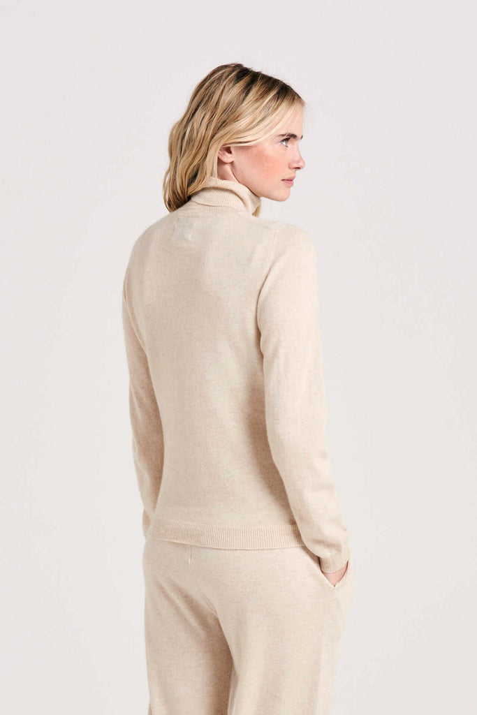 Blonde female model wearing Jumper1234 oatmeal cashmere roll neck facing away from the camera