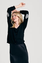 Blonde female model wearing Jumper1234 black cashmere cardigan with mid grey heart patch details on the elbows