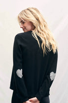 Blonde female model wearing Jumper1234 black cashmere cardigan with mid grey heart patch details on the elbows facing away from the camera