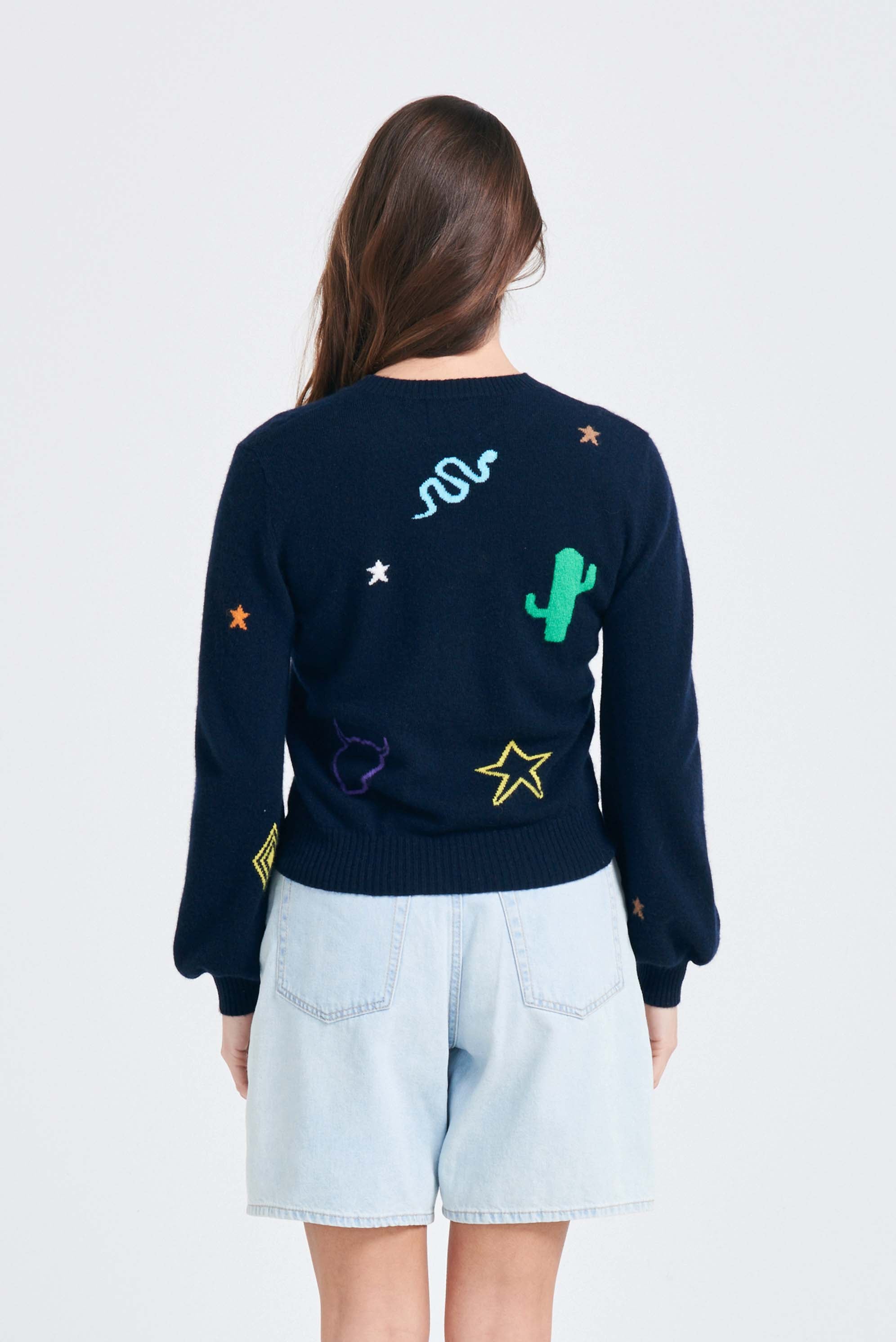 Brown haired female model wearing Jumper1234 Navy cashmere jumper with multi coloured cowboy icons back and front with slightly puff sleeves facing away from the camera