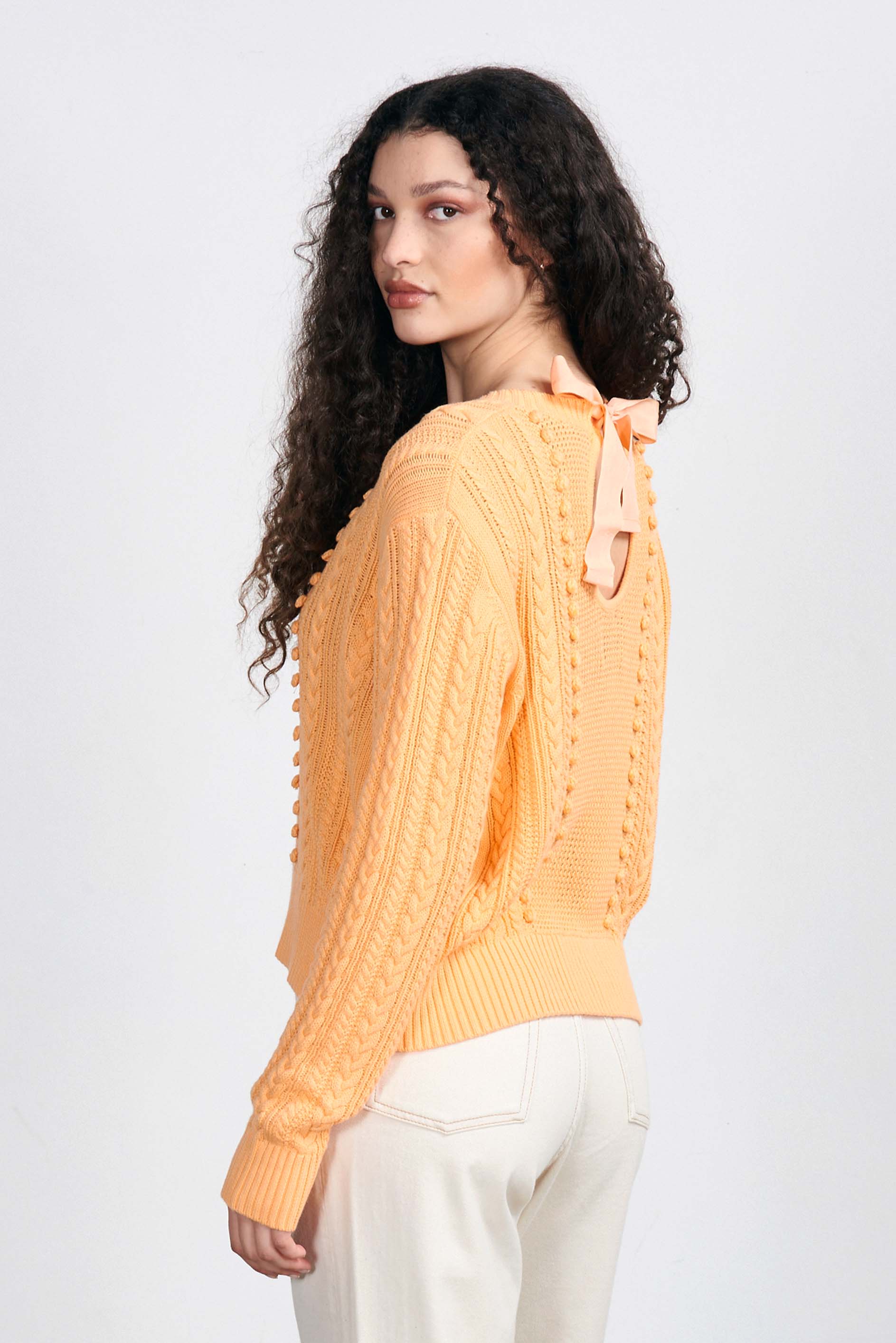 Brown haired female model wearing Jumper1234 Apricot cotton aran crew neck jumper with a ribbon tie back facing away from the camera