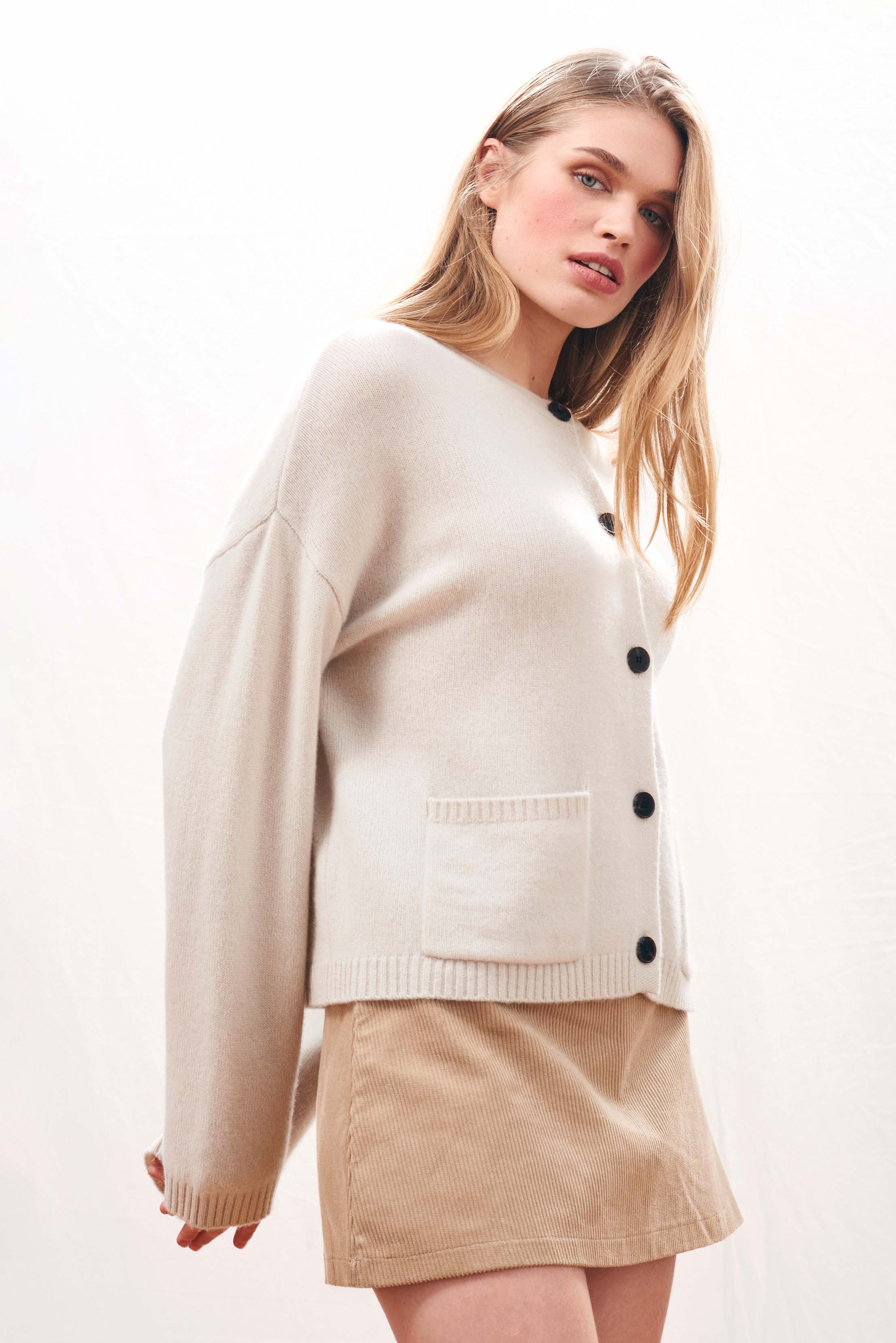 Female model wearing Jumper 1234 Heavier weight cashmere crew neck cardigan in stone. Oversize fit, two pockets and a fabulous cable detail at the back neck.