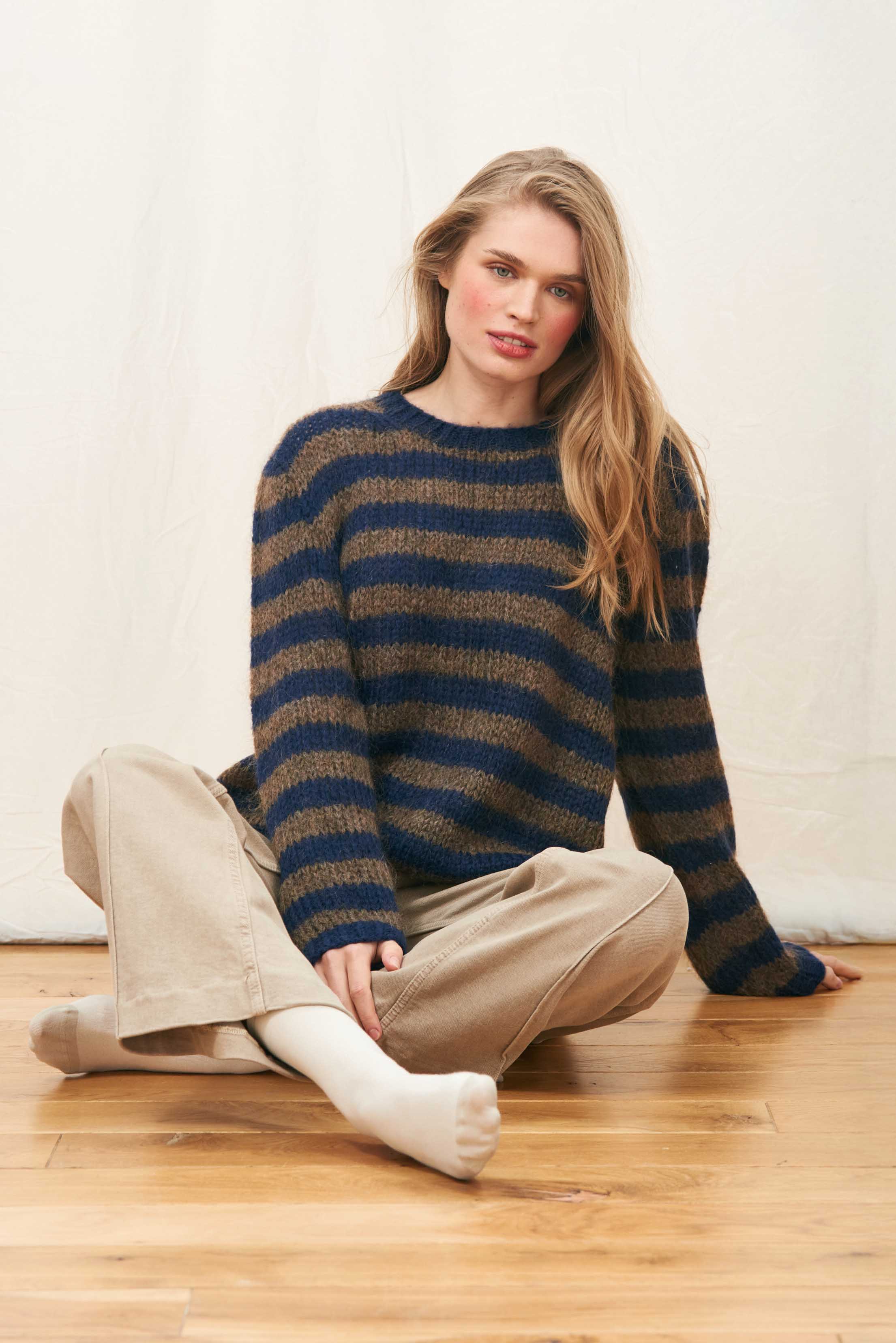 Female model sat on the floor wearing Jumper 1234 hand knitted mohair and wool blend hand knitted crew neck jumper in navy and chocolate stripes.