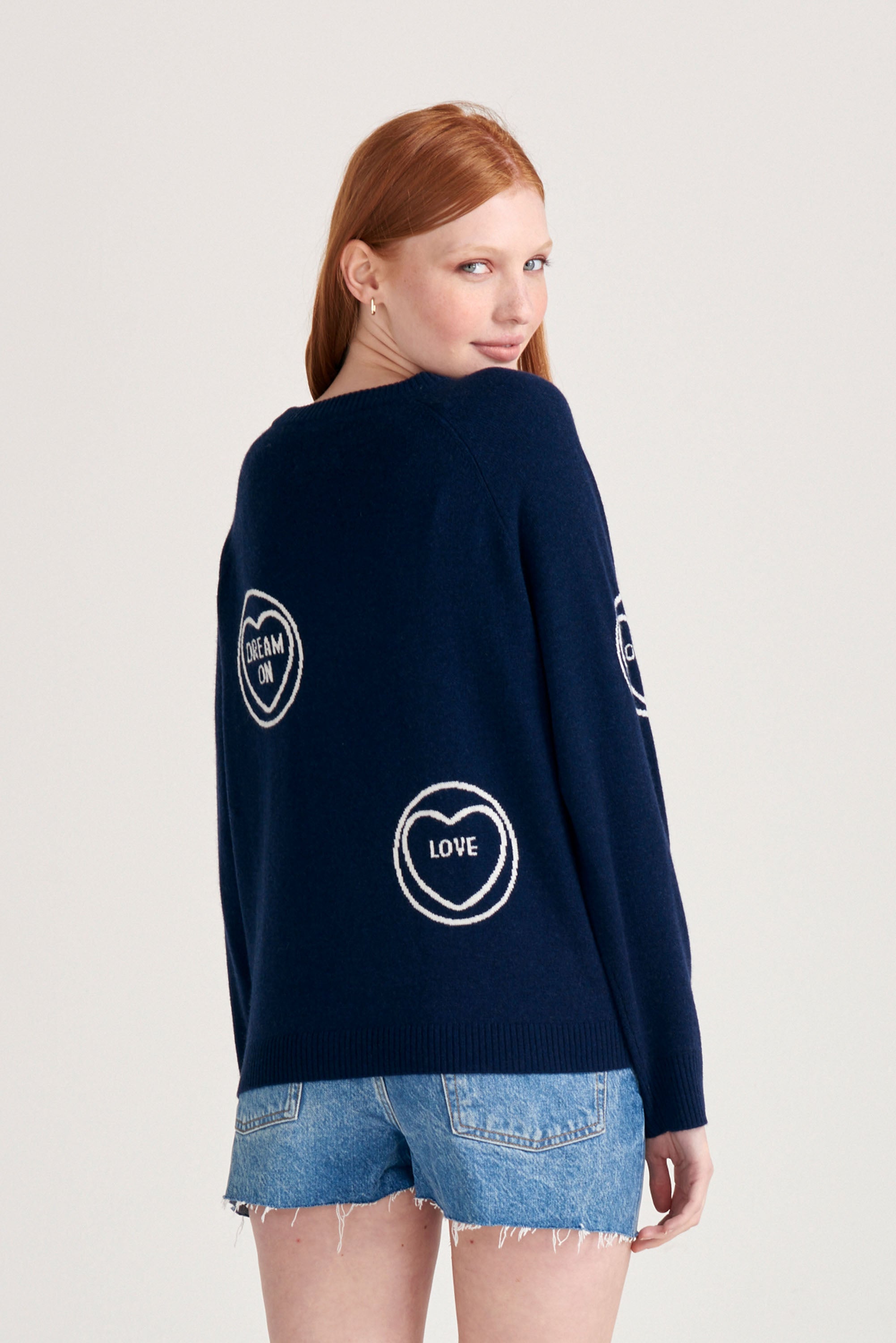 Red haired female model wearing Jumper1234 navy cashmere and wool mix crew neck sweatshirt style jumper with all over cream love hearts intarsia facing away from camera