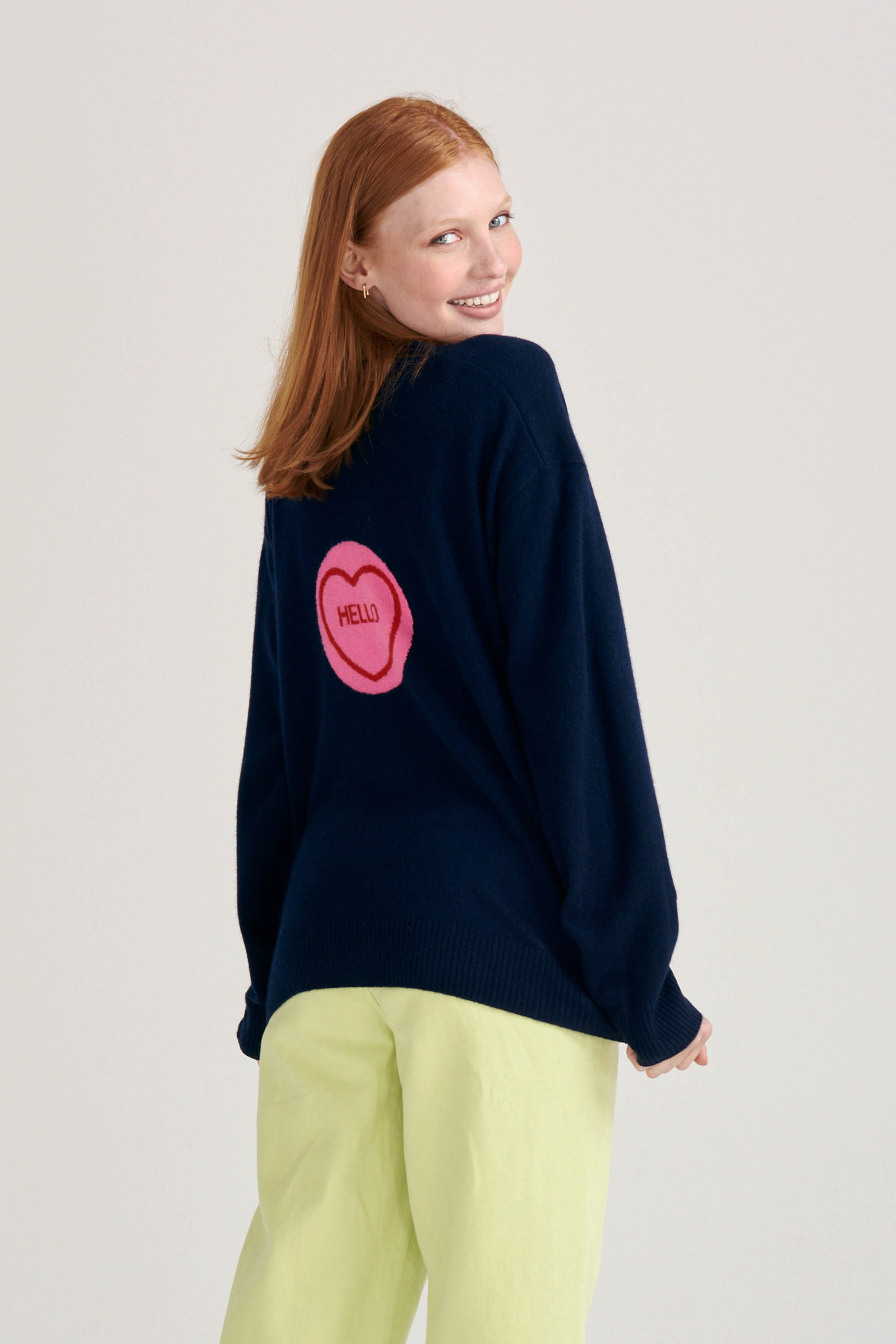 Red haired female model wearing Jumper1234 Navy cashmere vee neck cardigan with pink and red love heart 'hello' intarsia on the back facing away from the camera