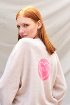 Red haired female model wearing Jumper1234 Oatmeal cashmere vee neck cardigan with hot pink and pale pink love heart 'hello' intarsia on the back facing away from the camera