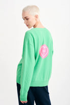 Blonde female model wearing Jumper1234 Bright green cashmere vee neck cardigan with pink love heart 'hello' intarsia on the back facing away from the camera