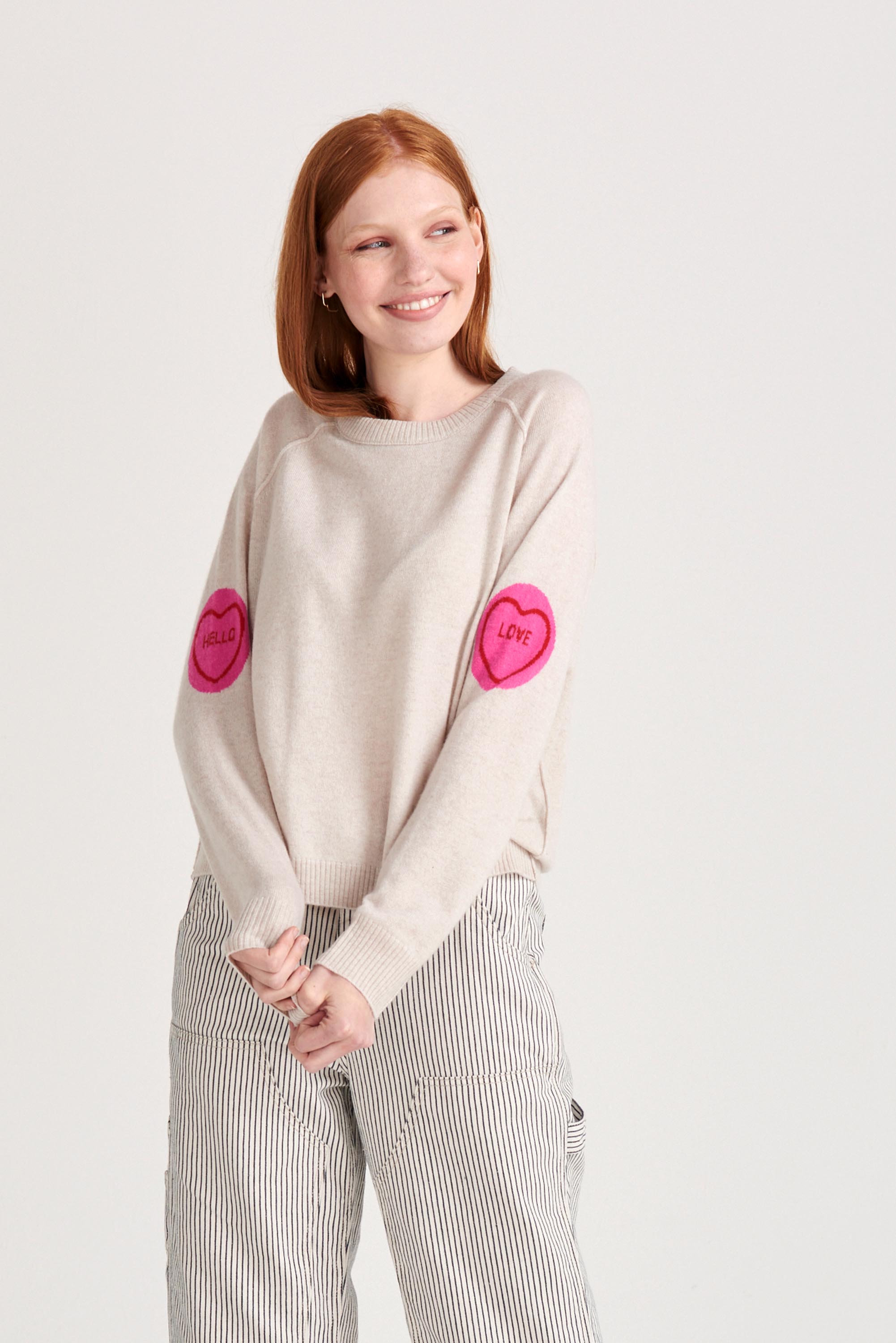 Red haired model wearing Jumper1234 Oatmeal cashmere and wool mix crew neck sweatshirt style jumper with all over pink and red love hearts intarsia on the sleeves