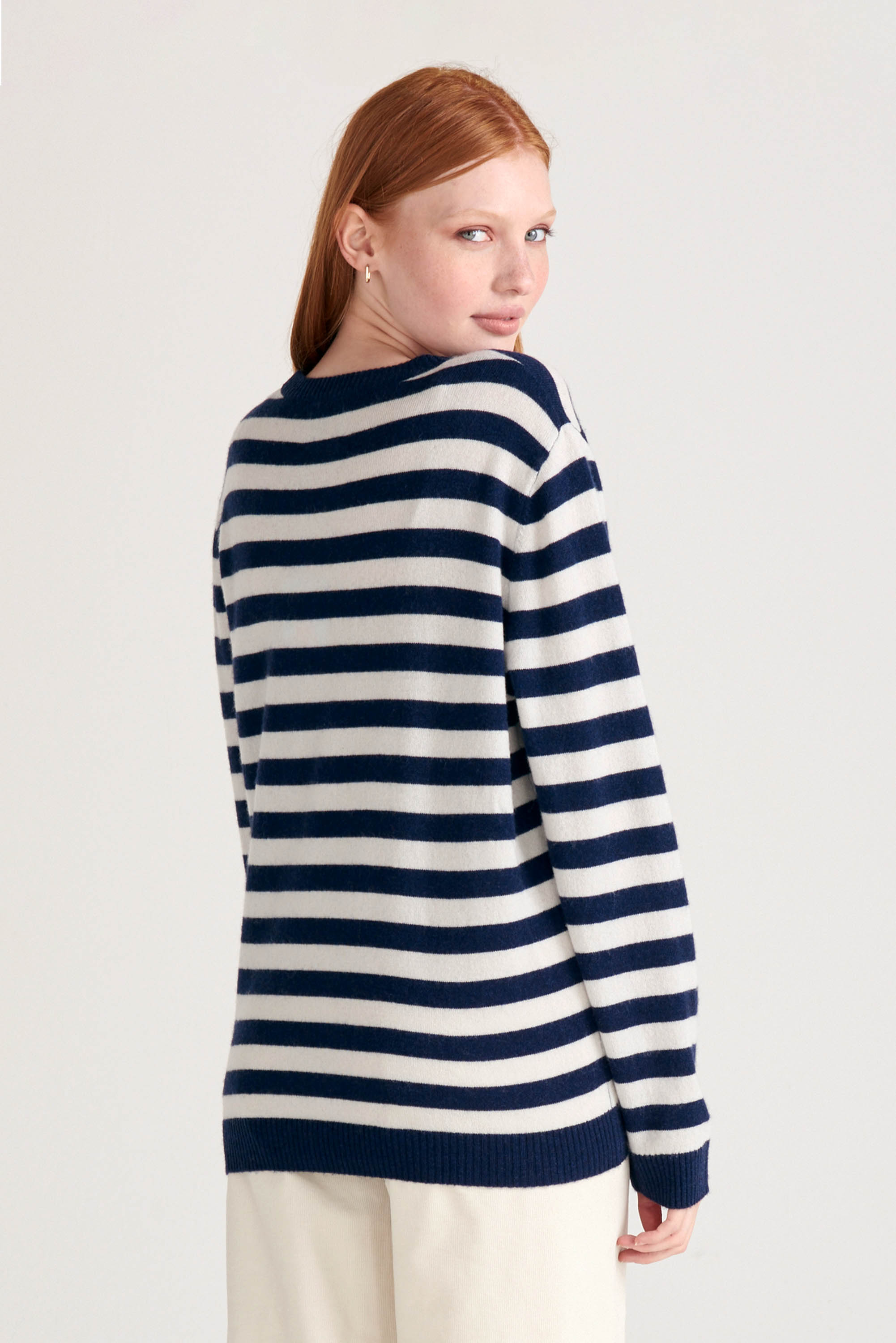 Red haired female model wearing Jumper1234 Navy and cream stripe cashmere and wool mix crew neck jumper with pink and red love heart intarsia facing away from the camera