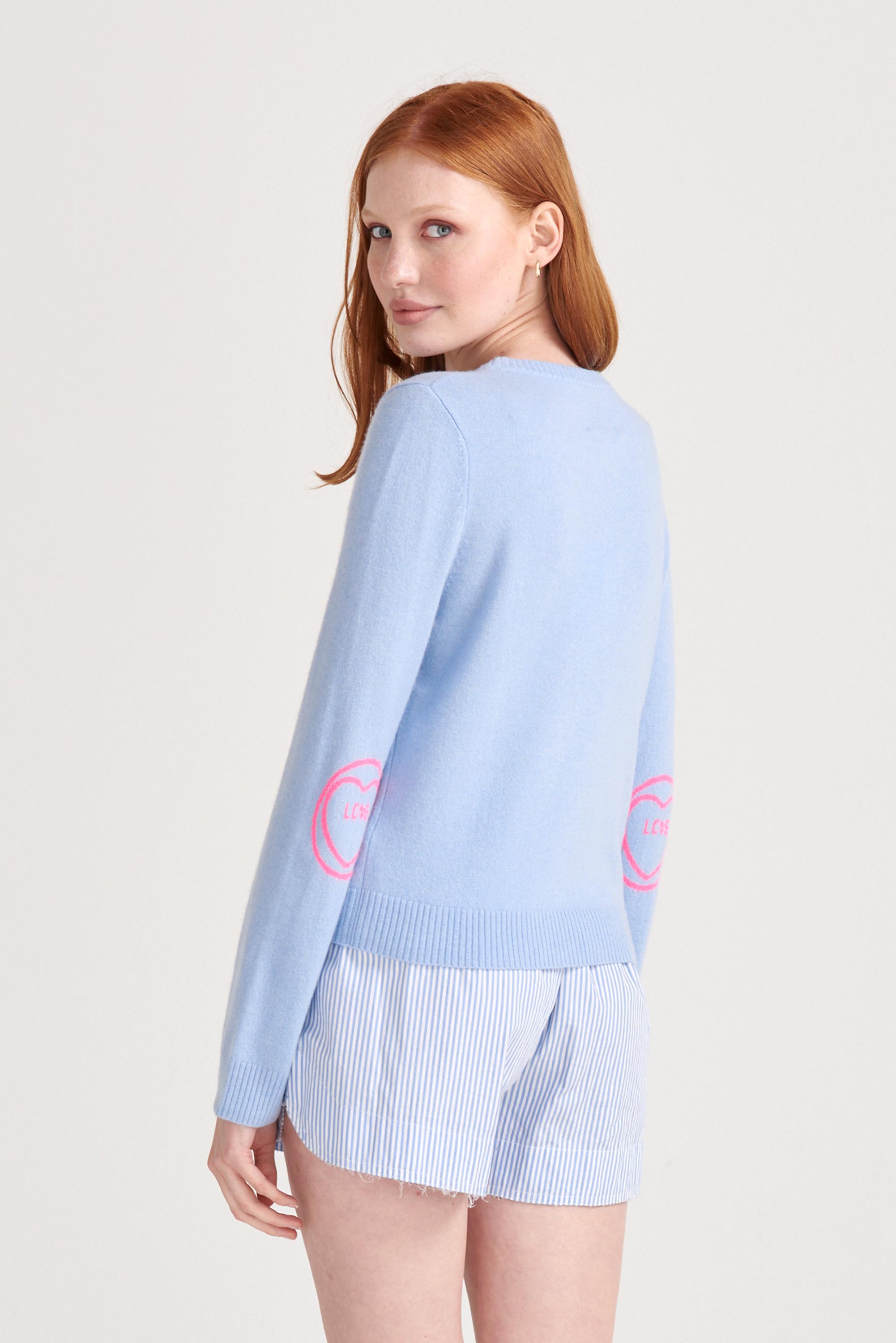 Red haired female model wearing Jumper1234 Pale blue cashmere crew neck jumper with neon pink love heart intarsia elbow patches facing away from the camera