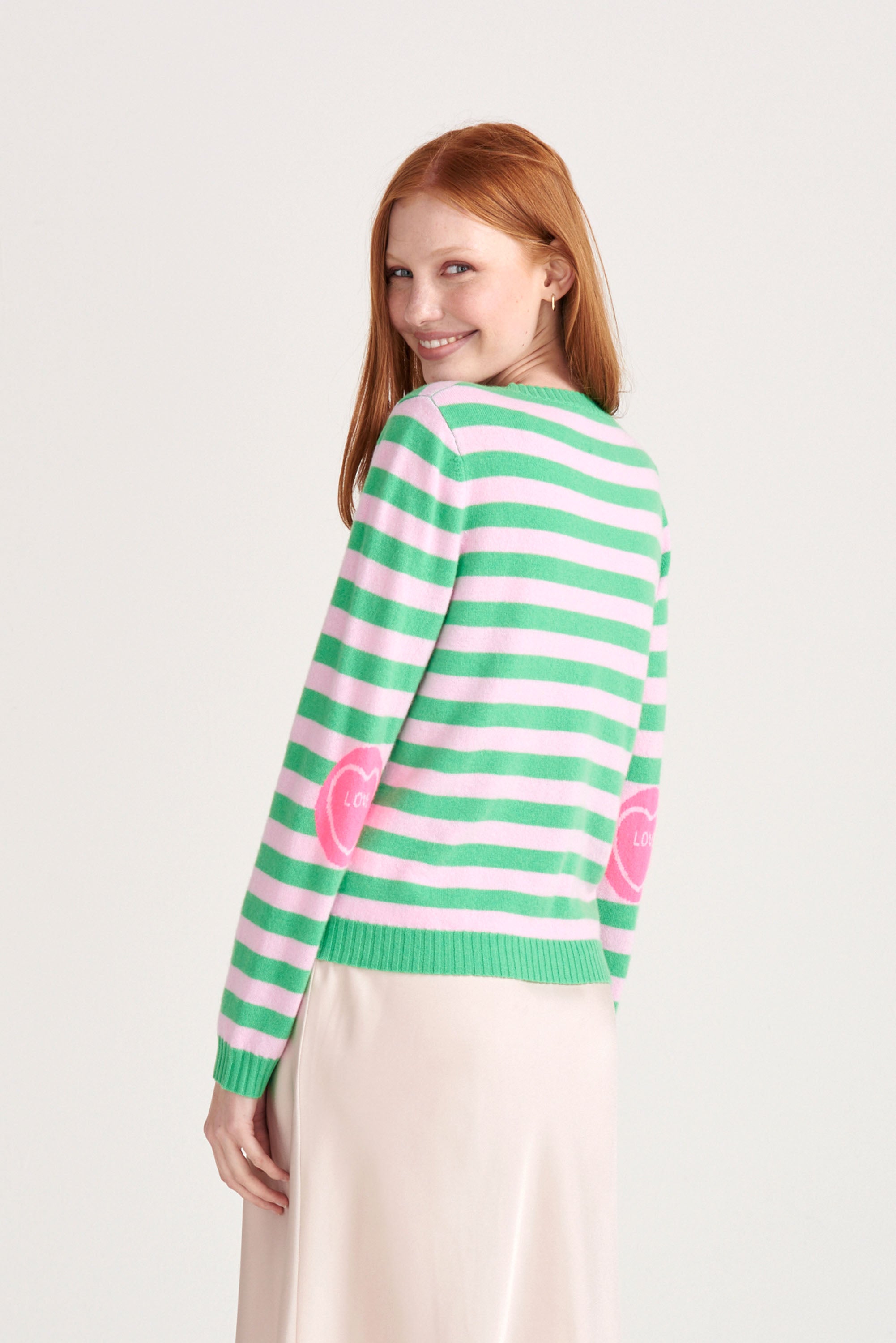 Red haired female model wearing Jumper1234 Bright green and pink stripe cashmere crew neck jumper with neon pink love heart intarsia elbow patches facing away from the camera