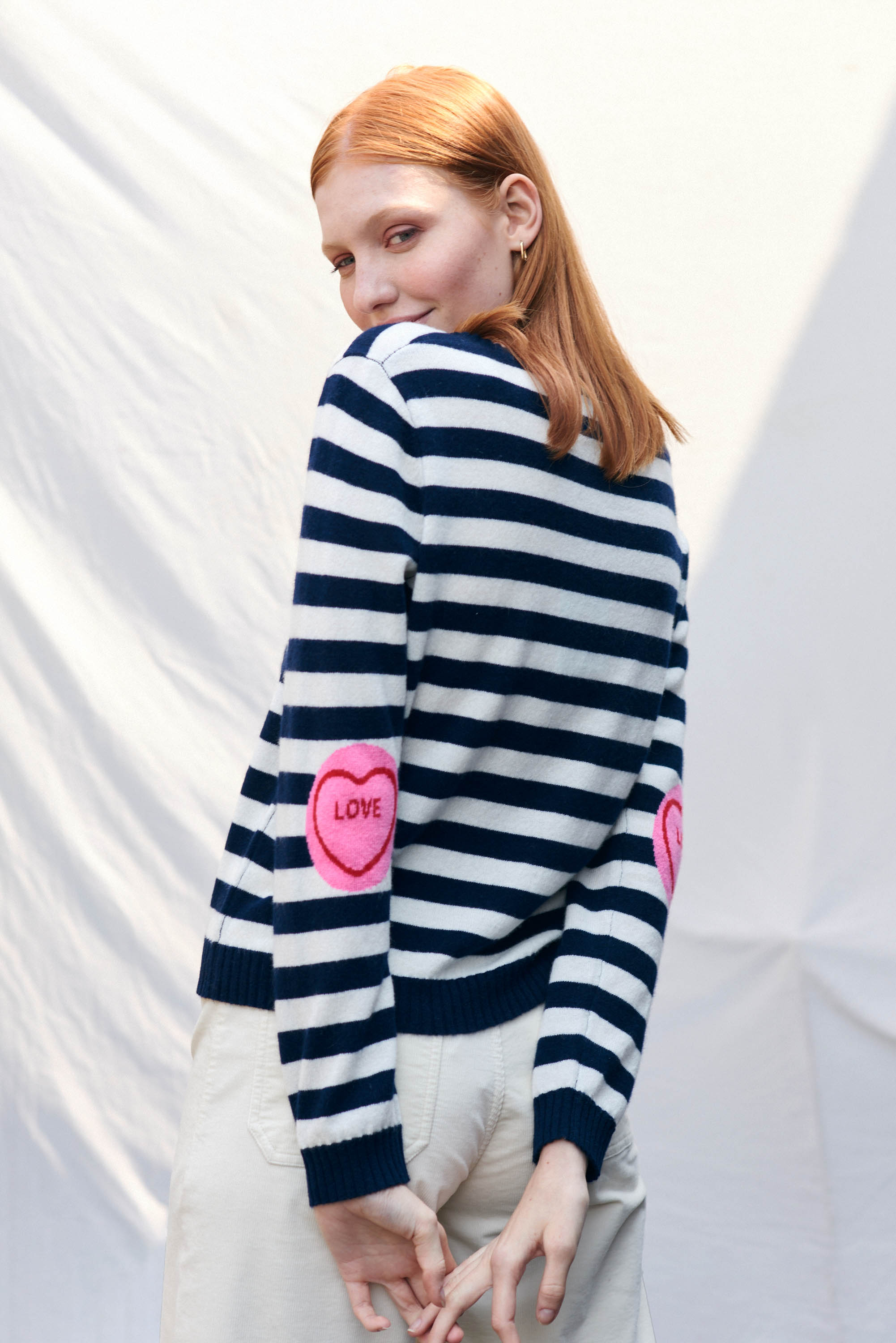 Red haired female model wearing Jumper1234 Navy and cream stripe cashmere crew neck jumper with pink love heart intarsia elbow patches facing way from the camera