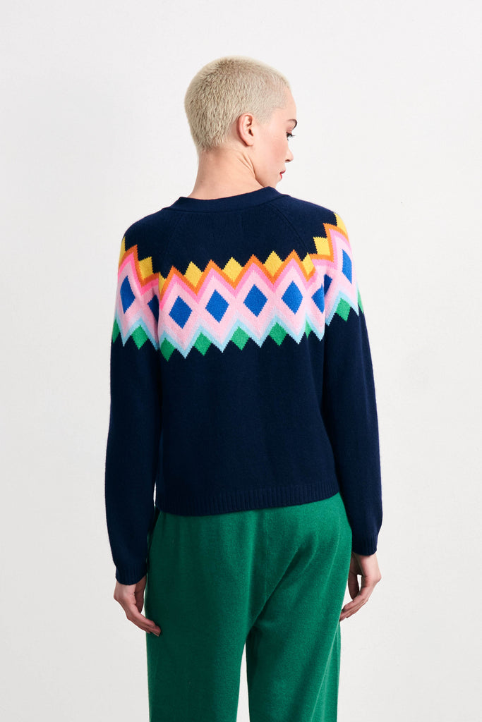Blonde female model wearing Jumper 1234 navy heavier weight cashmere vee neck, raglan sleeve cardigan with an intarsia Fairisle panel back and front in a fabulous combination of bright colours facing away from the camera