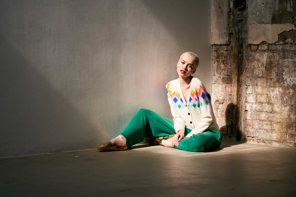 Blonde female model wearing Jumper 1234 cream heavier weight cashmere vee neck, raglan sleeve cardigan with an intarsia Fairisle panel back and front in a fabulous combination of bright colours sat on the floor