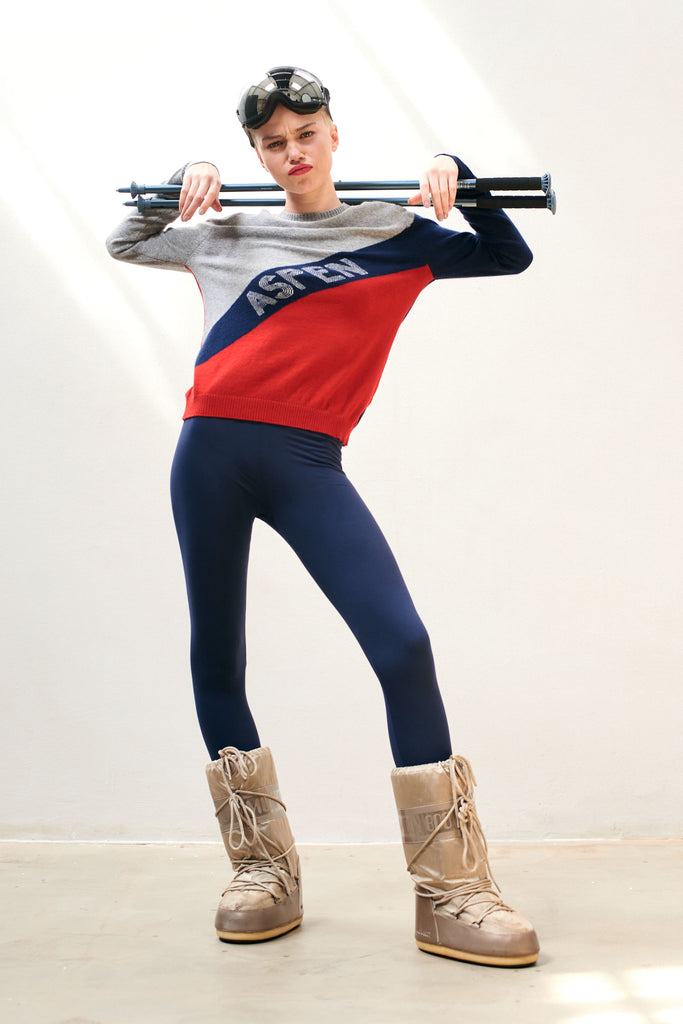 Blonde female model wearing Jumper1234 red, grey and navy colour block cashmere crew with cream "aspen" text diagonally across the chest holding ski poles