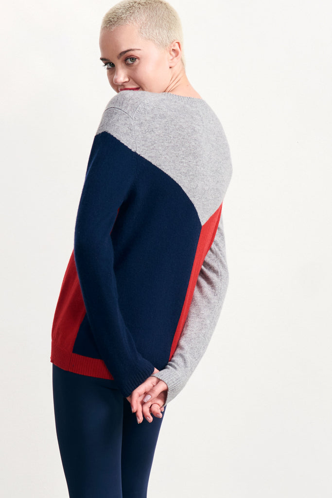 Blonde female model wearing Jumper1234 red, grey and navy colour block cashmere crew with cream "aspen" text diagonally across the chest facing away from the camera