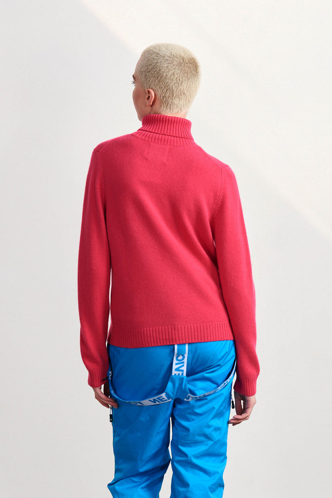 Blonde female model wearing Jumper 1234 crimson heavier weight roll neck cashmere and wool jumper with a cream panel and 'Vail' intarsia in crimson facing away from the camera