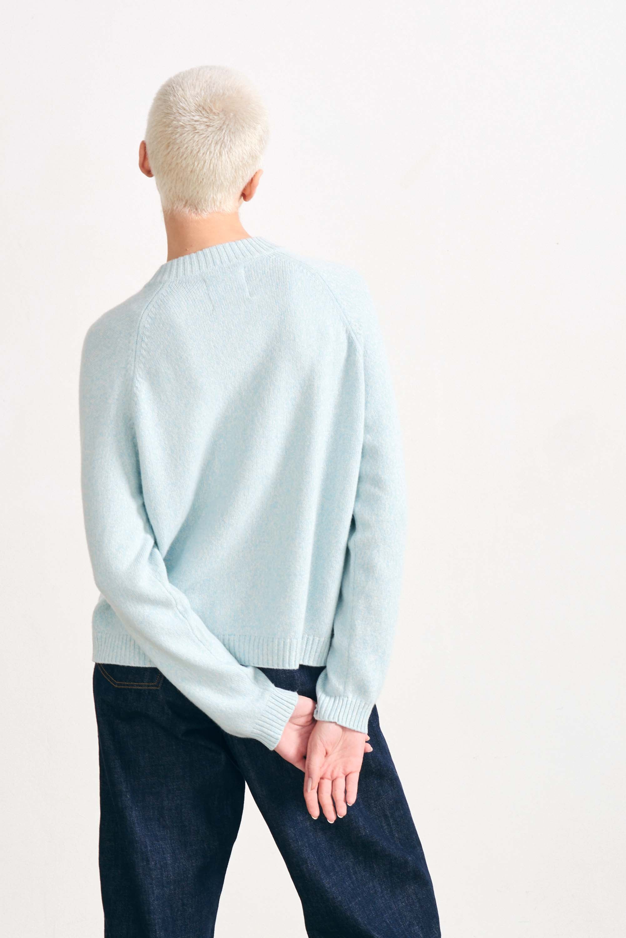 Blonde female model wearing Jumper 1234 cashmere and wool heavier weight round neck cardigan in ice blue marl facing away from the camera
