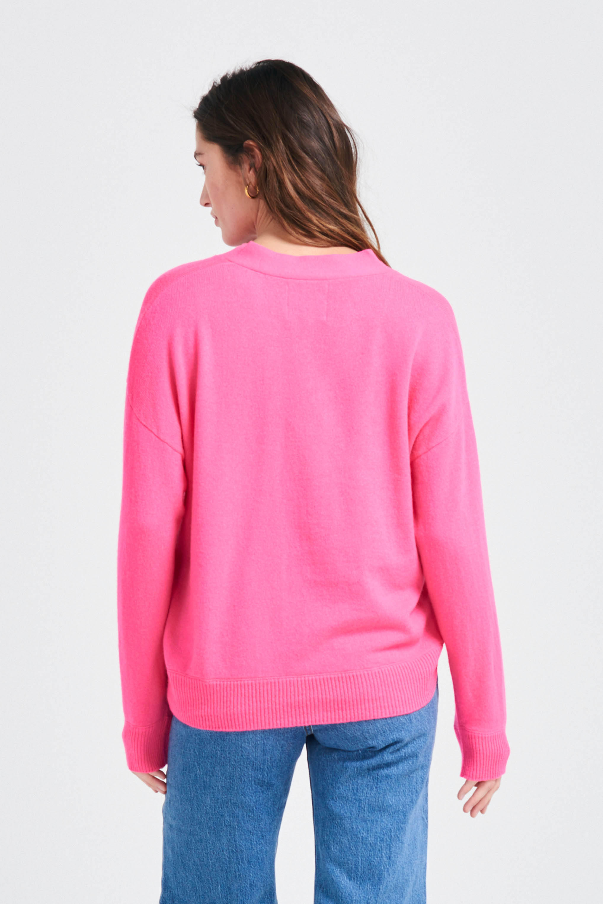 Brown haired female model wearing Jumper1234 Oversize cashmere vee neck cardigan in neon pink facing away from the camera