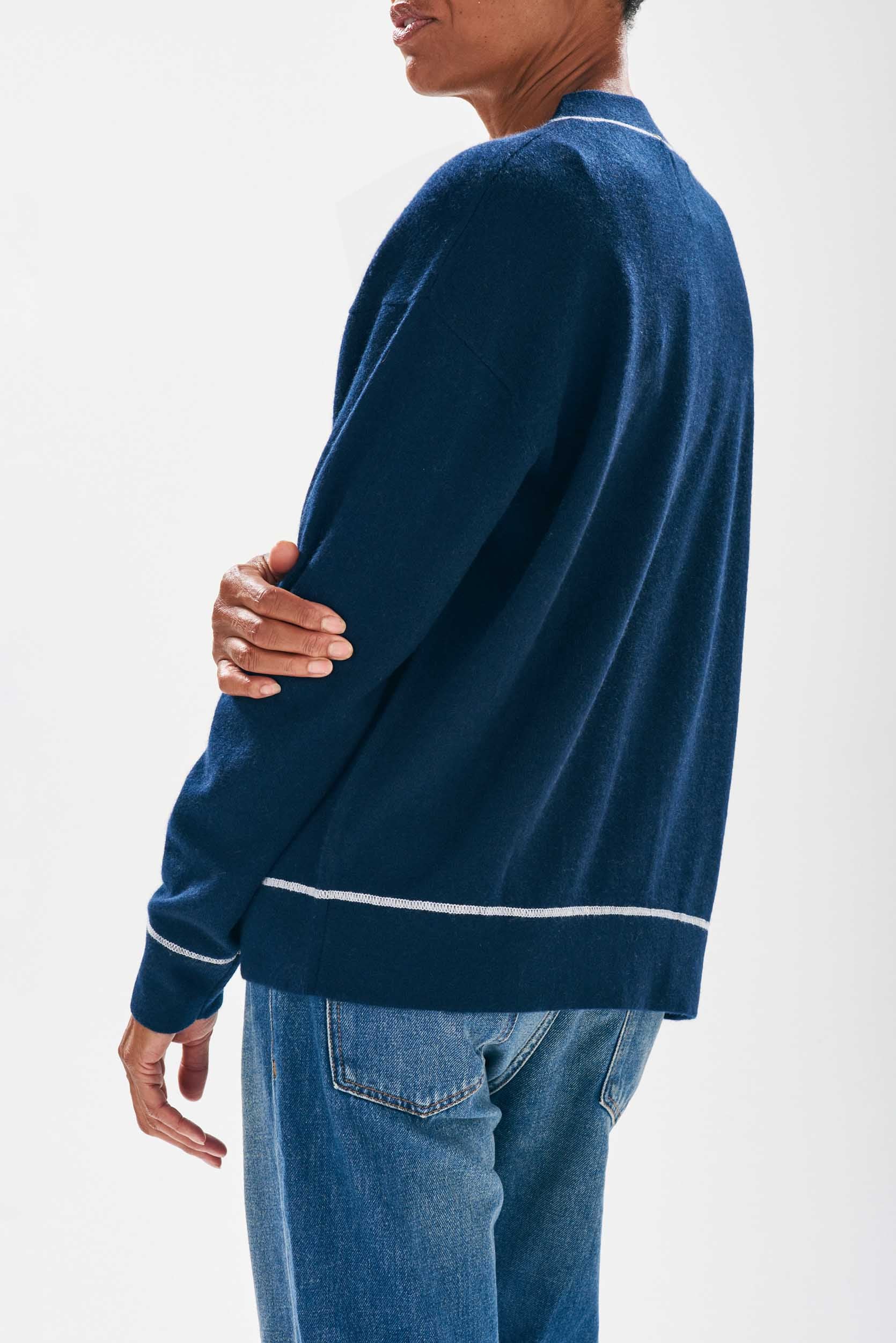 Model wearing Jumper1234 Navy boyfriend vee neck cashmere cardigan with cream top stitching detail and a pocket facing away from the camera