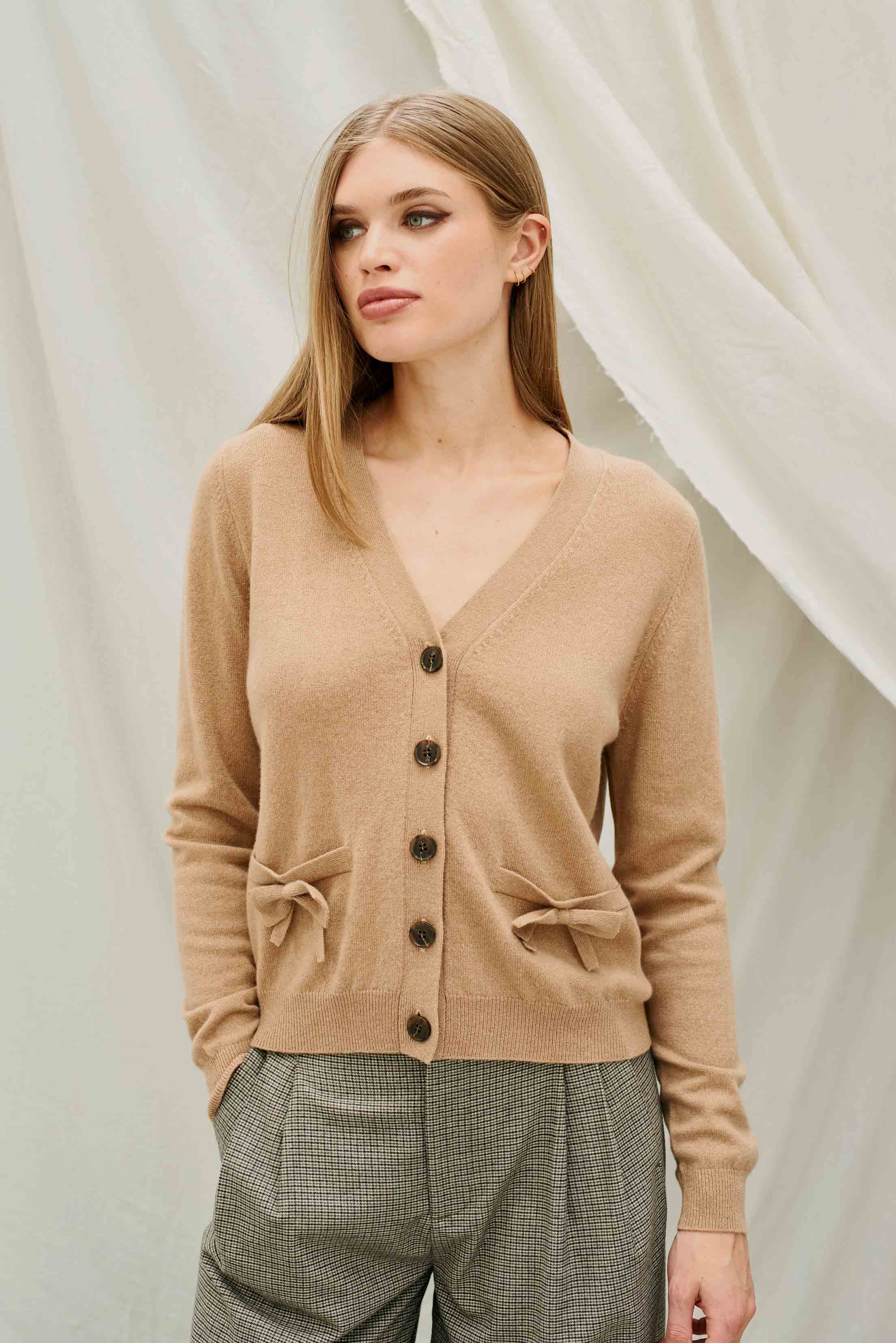 Female model wearing Jumper 1234 Camel vee neck cashmere cardigan with patch pockets with fabulous bows on.