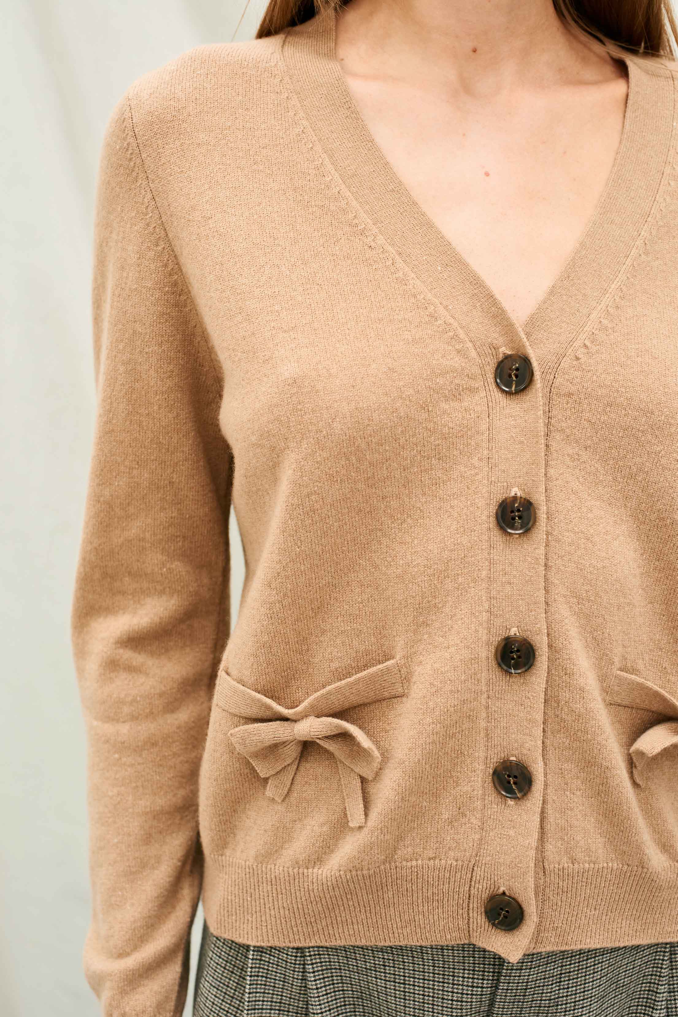 Female model wearing Jumper 1234 Camel vee neck cashmere cardigan with patch pockets with fabulous bows on. Close up
