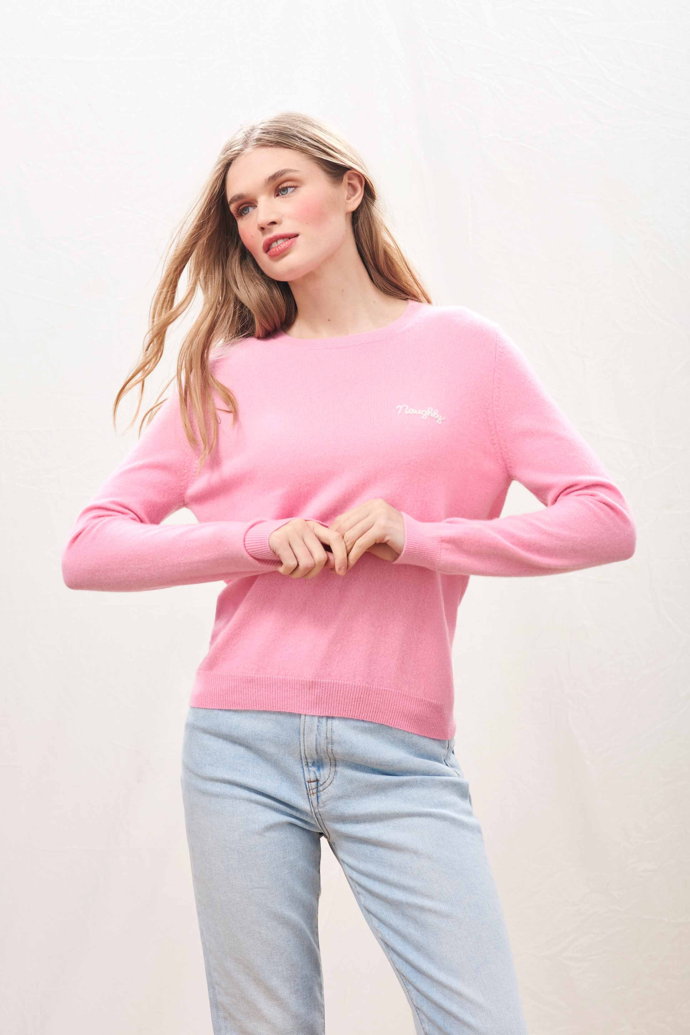 Female model wearing Jumper 1234 Candy cashmere crew neck jumper with cream 'naughty' chain stitch embroidery on the chest and 'but nice' on the back. 