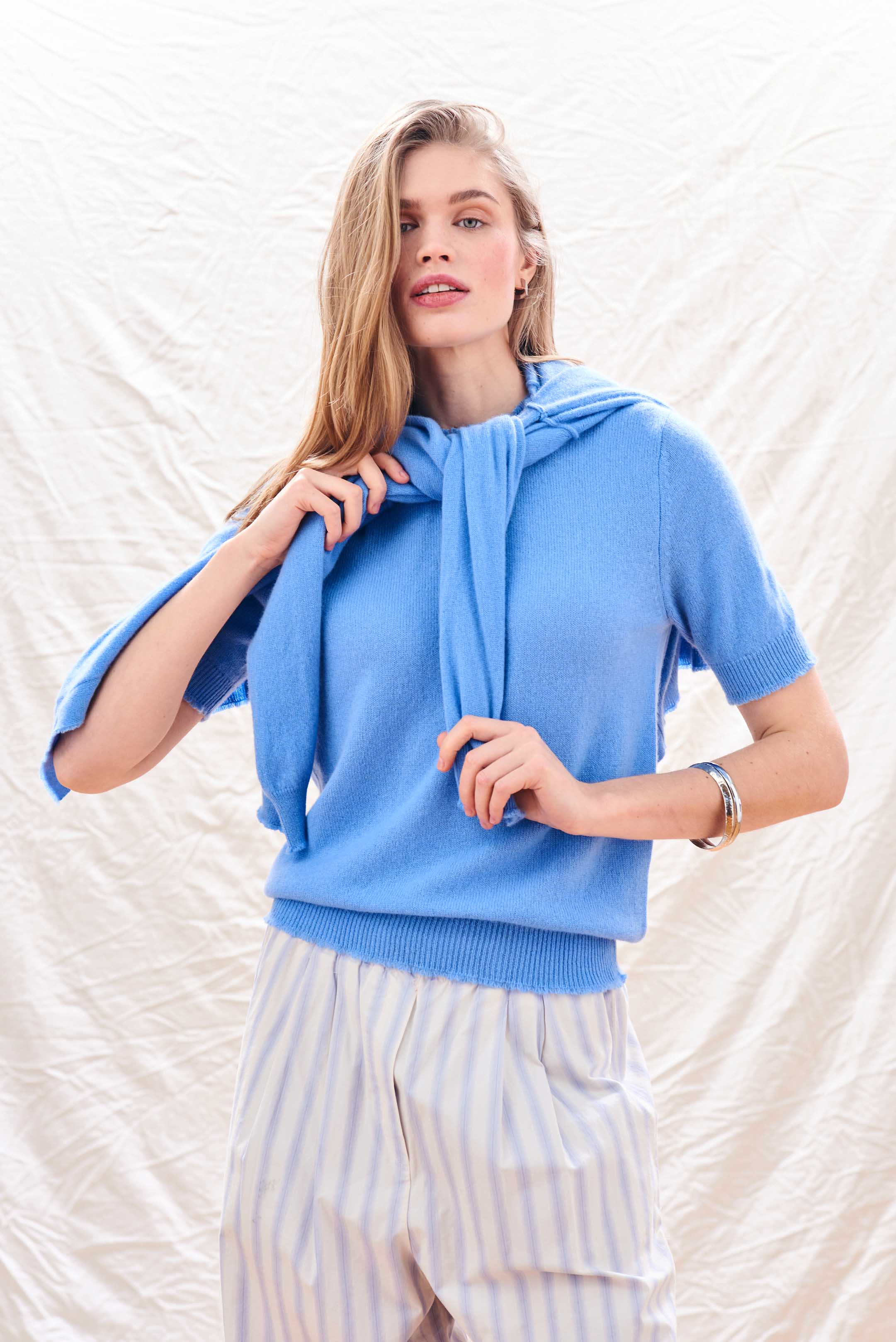 Blonde female model wearing Jumper 1234 Blue cashmere vee neck cardigan with 'distressed' ribs tied around her shoulders