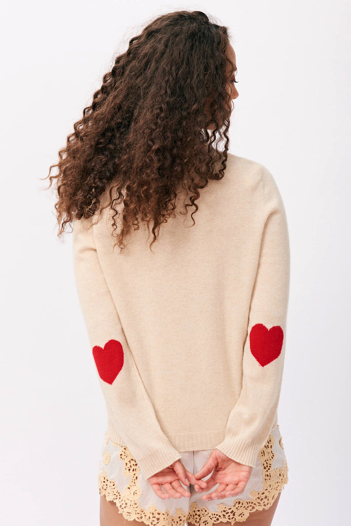 Brown haired female model wearing Jumper 1234 cashmere heart patch cardigan in oatmeal with red heart elbow patches facing away from the camera